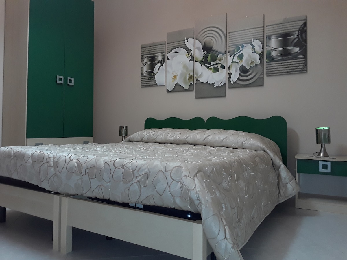A spacious bed with a light-colored quilt is positioned in the center of the room. Green elements, including a headboard and wardrobe, are present, complemented by a decorative wall art piece with white flowers. Two bedside tables with lamps offer additional lighting.