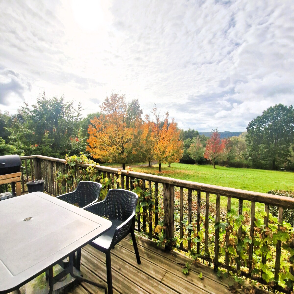 A wooden deck is shown with a table and two chairs facing an expansive view of vibrant trees showcasing autumn foliage. The sky is partially cloudy, allowing sunlight to illuminate the scene, enhancing the surrounding greenery.