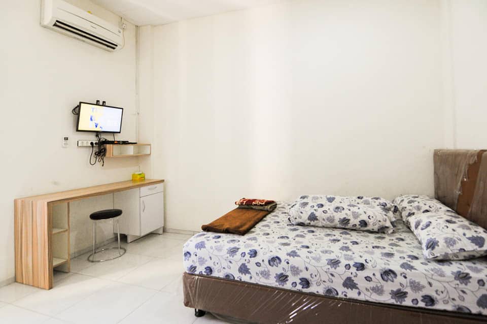 A simple room is equipped with a spacious bed featuring a floral-patterned mattress. A small wooden desk is positioned against the wall with a stool, providing a workspace. An air conditioning unit and a mounted television are present, while light-colored walls enhance the room's openness.