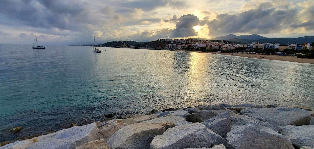 A calm shoreline view features smooth, large rocks in the foreground, transitioning to tranquil water. Sailboats are seen peacefully floating on the reflective surface, with a backdrop of rolling hills and town buildings under a softly lit sky.