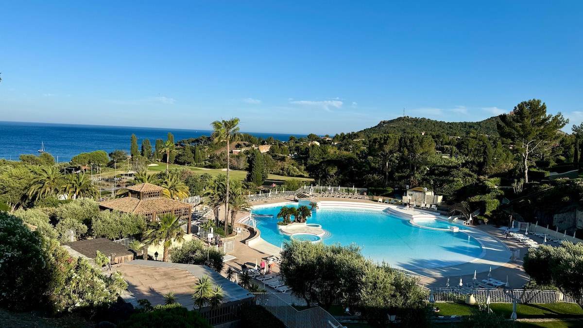 The image captures a serene view of a large swimming pool surrounded by lush greenery and palm trees. The pool reflects the clear blue sky and is situated against a backdrop of the Mediterranean Sea, creating a calm and inviting atmosphere.