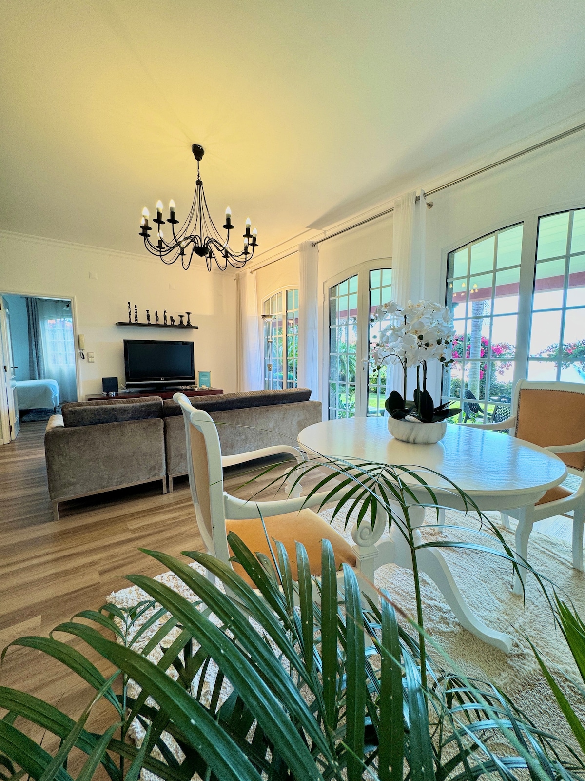 A welcoming living space features a stylish chandelier above a round dining table, surrounded by light-colored chairs. Large glass doors provide views of the outdoor area, while a soft rug and indoor plants add to the serene ambiance.