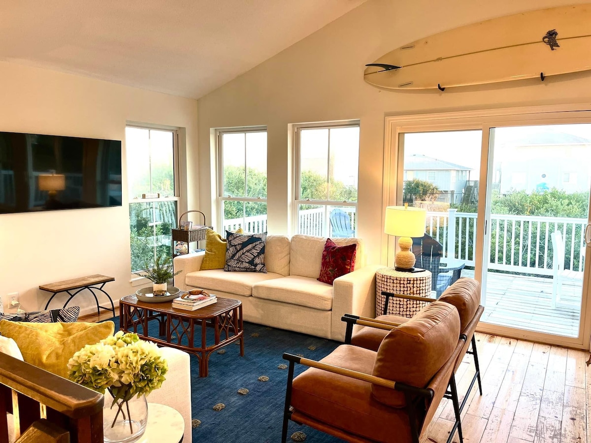 A bright living area features large windows offering ample natural light. A light-colored sectional sofa is adorned with decorative pillows. A coffee table rests on a blue rug, while a vintage-style chair and a surfboard hang above, adding a touch of coastal charm.