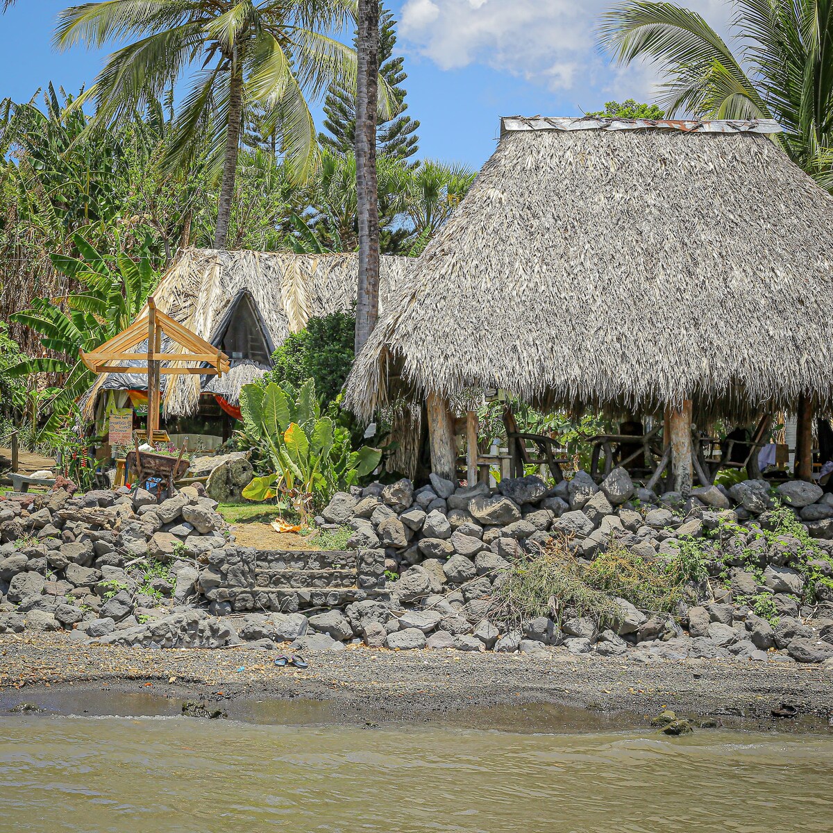 A thatched-roof cabin is situated near a rocky shoreline, surrounded by tropical plants and palm trees. A wooden gazebo with a simple design is also visible. The scene captures the natural beauty of the area by the lake, creating a serene environment.