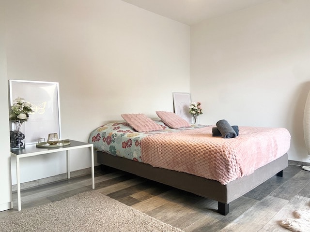 A spacious sleeping area features a large bed with a soft pink blanket and floral bedding. A small side table holds decorative items, including flowers in a vase. Natural light enters through two adjacent sources, illuminating the neutral-toned walls and flooring.