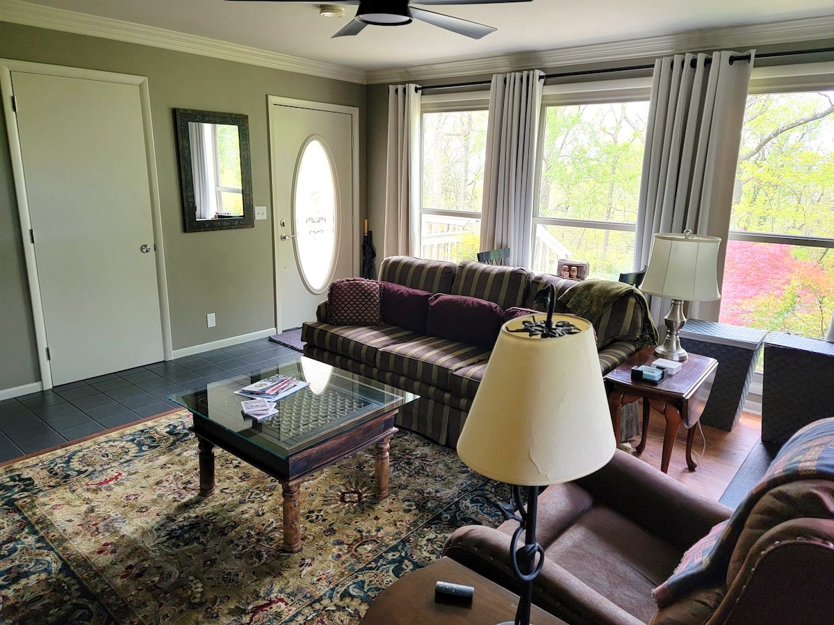A spacious living area features a combination of dark wood and brown furniture, including a large sofa with patterned upholstery and armchairs. Natural light filters through the large windows, revealing views of the surrounding greenery. A decorative area rug adds warmth to the tiled floor, while a round entry door enhances the inviting atmosphere.