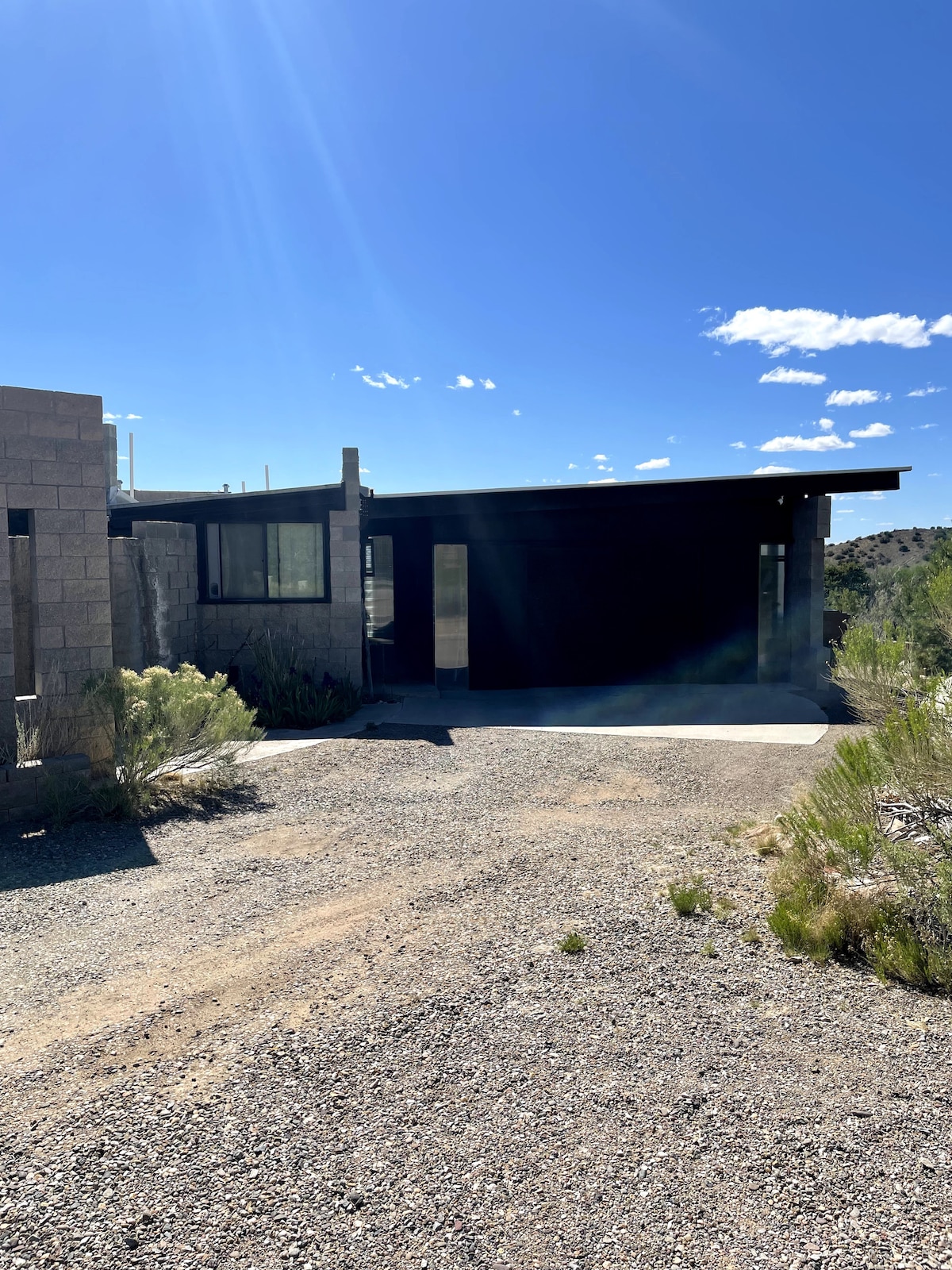A modern dwelling is visible, featuring a combination of stone and wood elements. The building has expansive windows allowing natural light, while a covered porch area extends over the entrance. Surrounding vegetation includes low shrubs and desert plants, set against a clear blue sky.