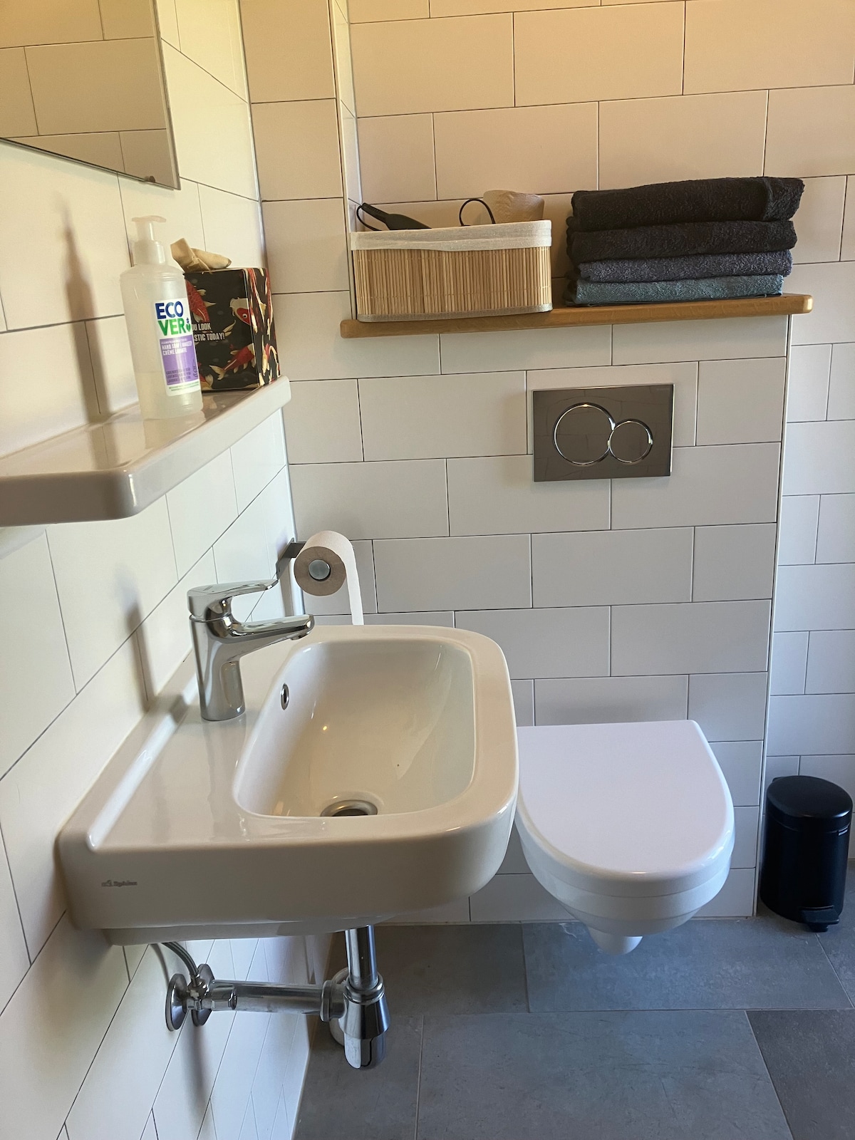 A modern bathroom features a compact white sink with a chrome faucet, positioned beside a wall-mounted toilet. The wall is lined with light-colored tiles, while a shelf above holds neatly stacked dark towels and a storage basket. Personal care items are visible on the sink.
