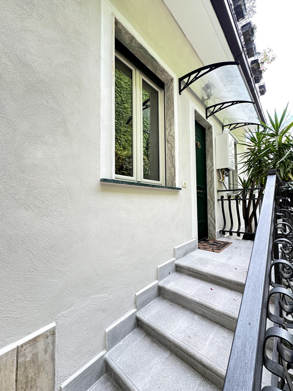 A private entrance is seen, featuring a set of light-colored stairs leading up to a green door. Decorative awning shades the entryway, and greenery surrounds the area, adding a touch of nature.