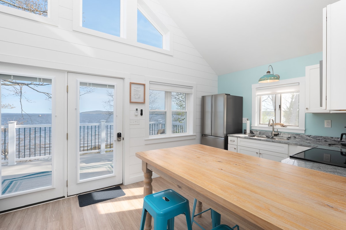 The kitchen features large windows that offer a view of the ocean, allowing natural light to fill the space. A wooden dining table, complemented by teal stools, is positioned centrally. Stainless steel appliances are showcased against a light-colored backdrop.