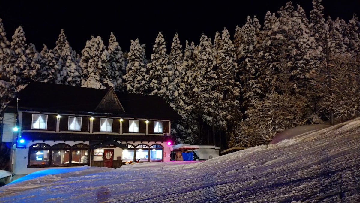 The lodge is nestled among tall, snow-covered trees, creating a serene winter atmosphere. The building features multiple illuminated windows, with soft light spilling out, contrasting against the snowy landscape. A gentle slope leads towards the entrance, inviting guests to explore.