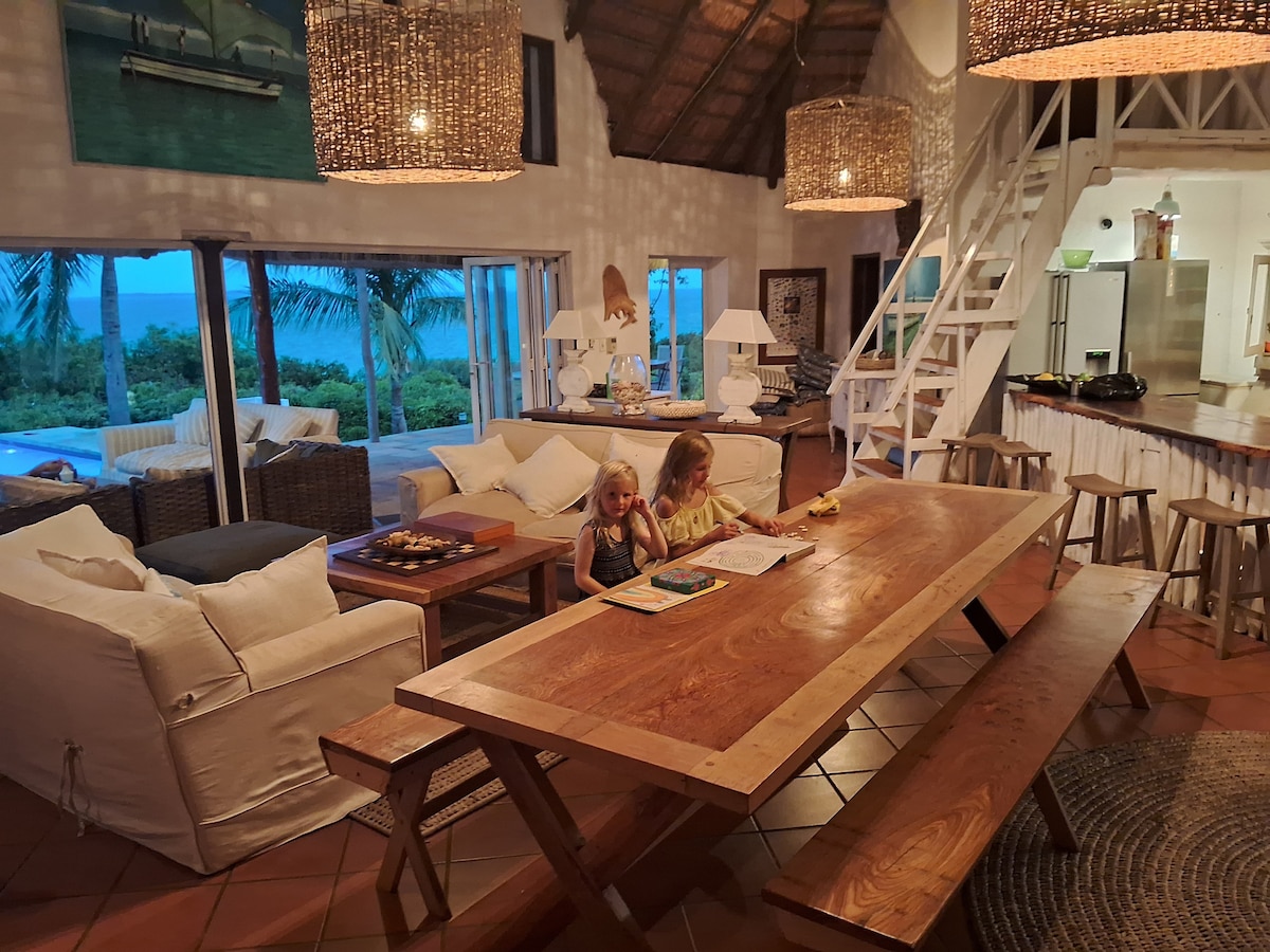 The open-plan living area features a wooden dining table surrounded by natural light, with a cozy seating arrangement consisting of large white sofas. Tropical views of palm trees and the ocean can be seen through expansive glass doors, enhancing the spacious feel of the room.