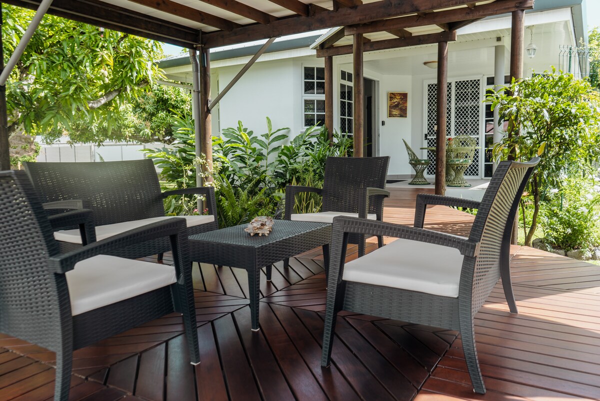 A covered deck area features four outdoor chairs arranged around a small table. Lush greenery surrounds the space, complementing the serene view of the nearby bungalow. Natural light filters through the open structure, creating a relaxing outdoor environment.