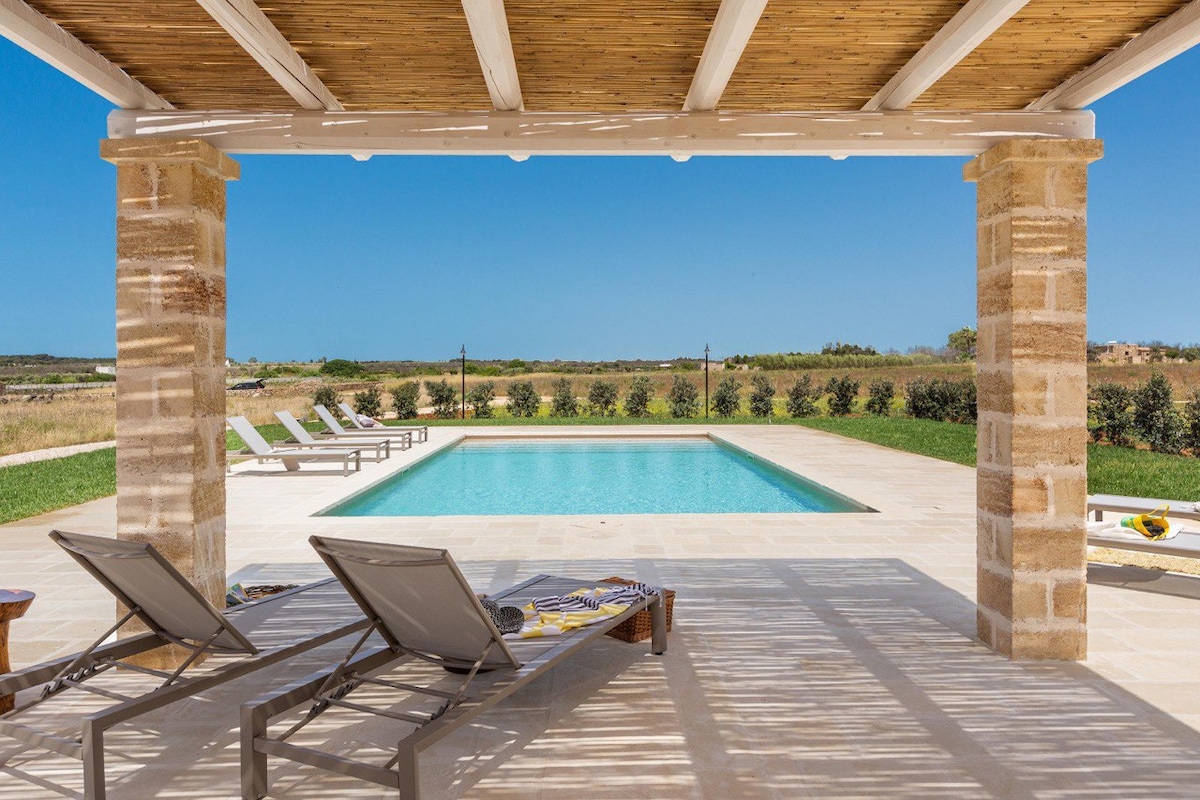 A covered terrace provides a shaded view of a serene swimming pool, surrounded by a spacious sun deck with loungers. An open landscape extends in the background, with greenery and clear blue skies enhancing the outdoor leisure area.