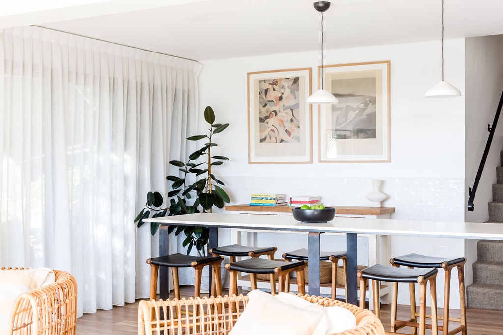 A bright dining area is presented, featuring a long table surrounded by six wooden chairs. Above, two pendant lights hang gently. A large potted plant stands beside the table, and soft white curtains filter natural light from the nearby window.