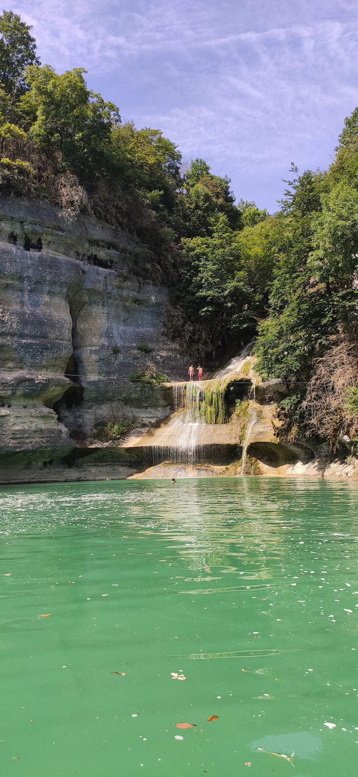 A natural landscape features a waterfall cascading down rocky cliffs surrounded by lush greenery. The clear turquoise water reflects the vibrant colors of the scene. Two figures are positioned at the top of the waterfall, emphasizing the scale of the serene environment.