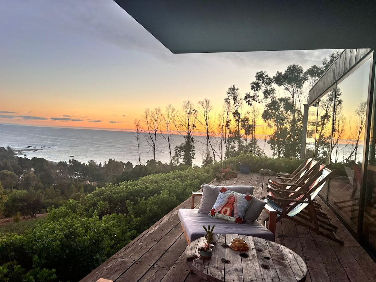 An outdoor terrace is presented, featuring a comfortable seating area that overlooks the ocean. Wooden lounge chairs are positioned alongside a low coffee table adorned with decorative items. The horizon displays a warm sunset, casting soft light across the tranquil landscape dotted with lush greenery.