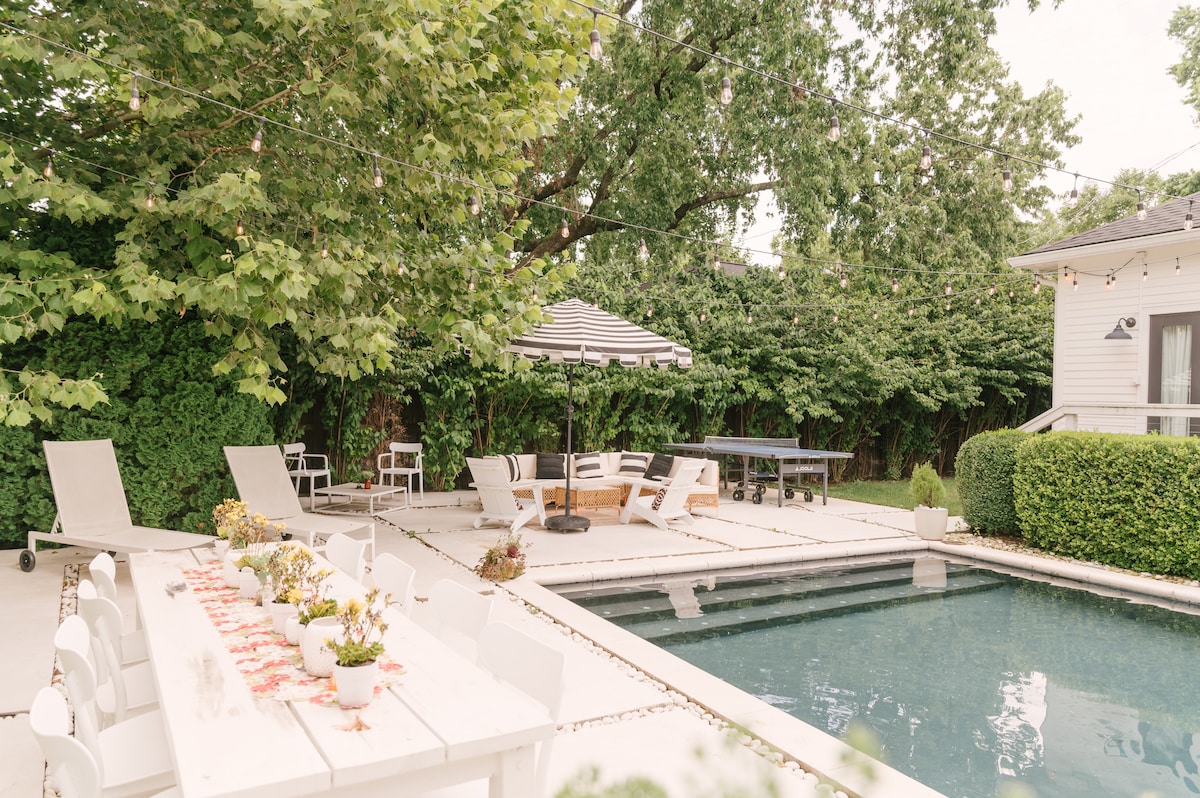 An outdoor space featuring a heated saltwater pool, surrounded by lush greenery. A long dining table is set with potted plants, while lounge chairs are positioned nearby. A striped umbrella provides shade over a seating area, creating a relaxed atmosphere for gatherings.