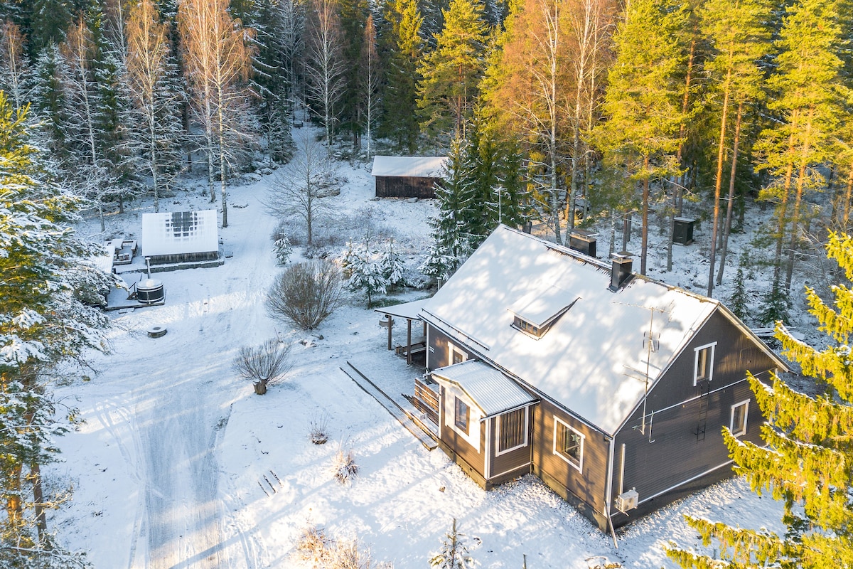 An aerial view of a winter landscape reveals a spacious wooden house surrounded by snow-covered trees. A pathway leads through the snowy ground, connecting to various outbuildings, including a sauna and storage shed visible in the background.