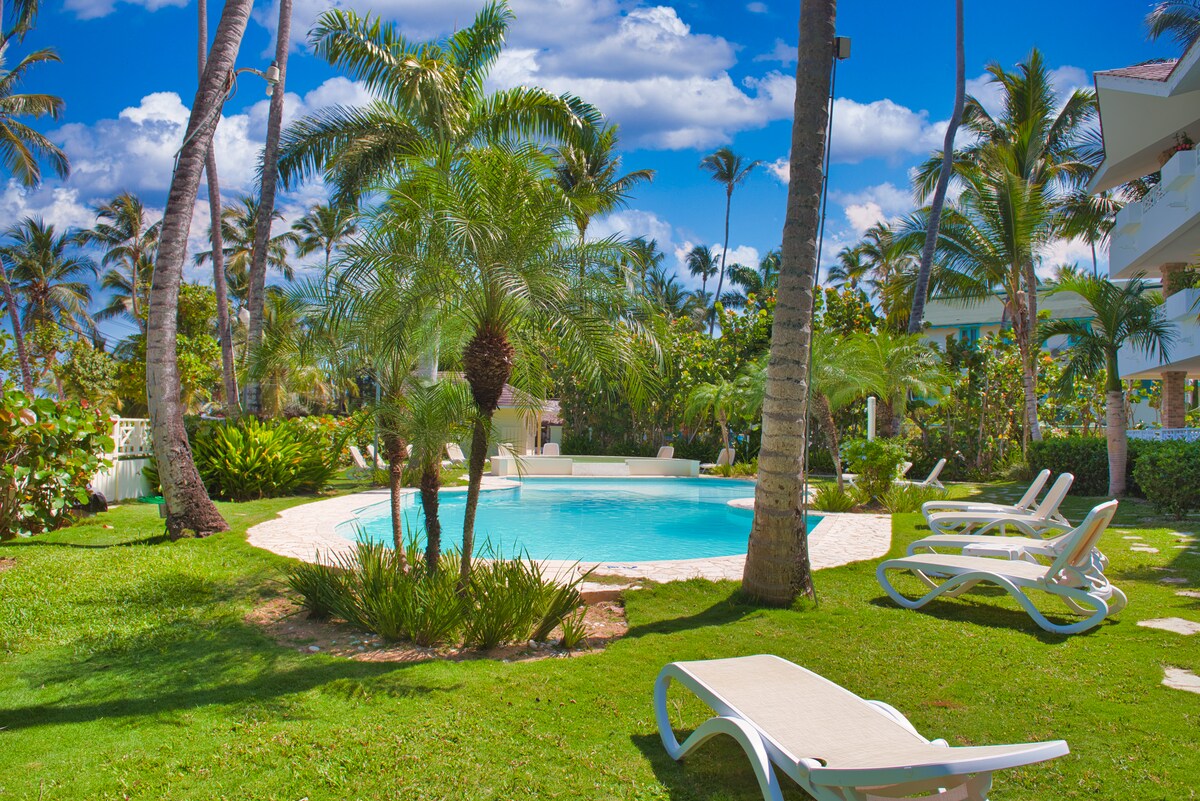 A serene outdoor area features a circular swimming pool surrounded by lush tropical greenery. White lounge chairs are positioned on the grass, providing a relaxing spot for sunbathing. Tall palm trees sway gently in the background under a bright blue sky with scattered clouds.