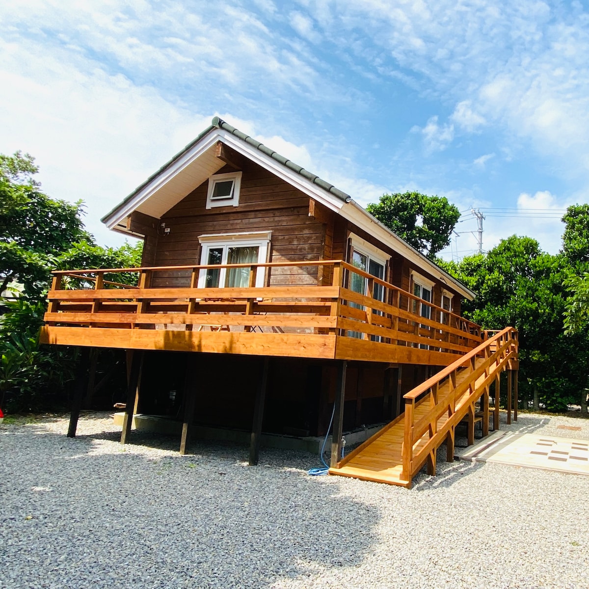 A wooden log house is elevated on stilts, surrounded by greenery. A spacious wooden deck with railings wraps around the front, offering outdoor seating. Steps lead up to the entrance, and a gravel area provides additional parking space beneath clear blue skies.