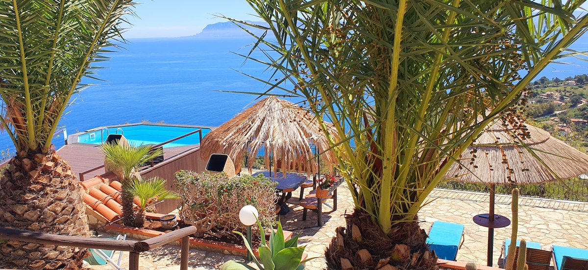 An outdoor area is framed by lush palm trees, featuring a seating arrangement with a table and chairs. A thatched-roof gazebo provides shade, while a pool can be seen in the distance, overlooking the expansive blue sea and horizon.