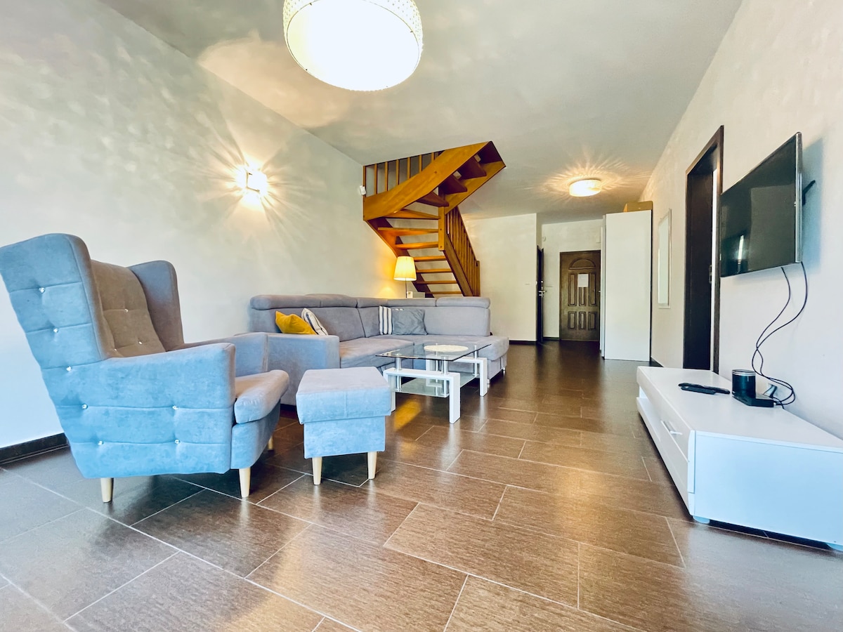 A comfortable living area features a gray sectional sofa and armchair, complemented by a coffee table. A flat-screen TV is mounted on a white media console. Natural light fills the space, highlighting the staircase leading to an upper level.