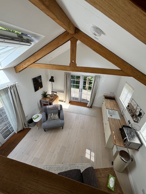 An open-plan living area is highlighted by wooden beams that create a spacious feel. Natural light floods in through large windows, illuminating the light wood flooring. A small dining table and kitchen area are visible, alongside a comfortable seating arrangement by the door leading to the garden.
