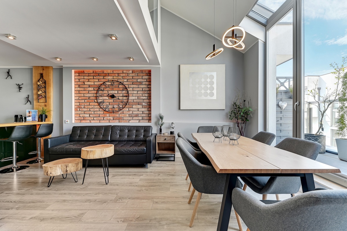 An open living space features a brick accent wall, a sleek black sofa, and a wooden dining table surrounded by stylish chairs. Large windows allow natural light to brighten the area, while decorative plants enhance the ambiance. A combination of modern lighting fixtures adds subtle illumination.