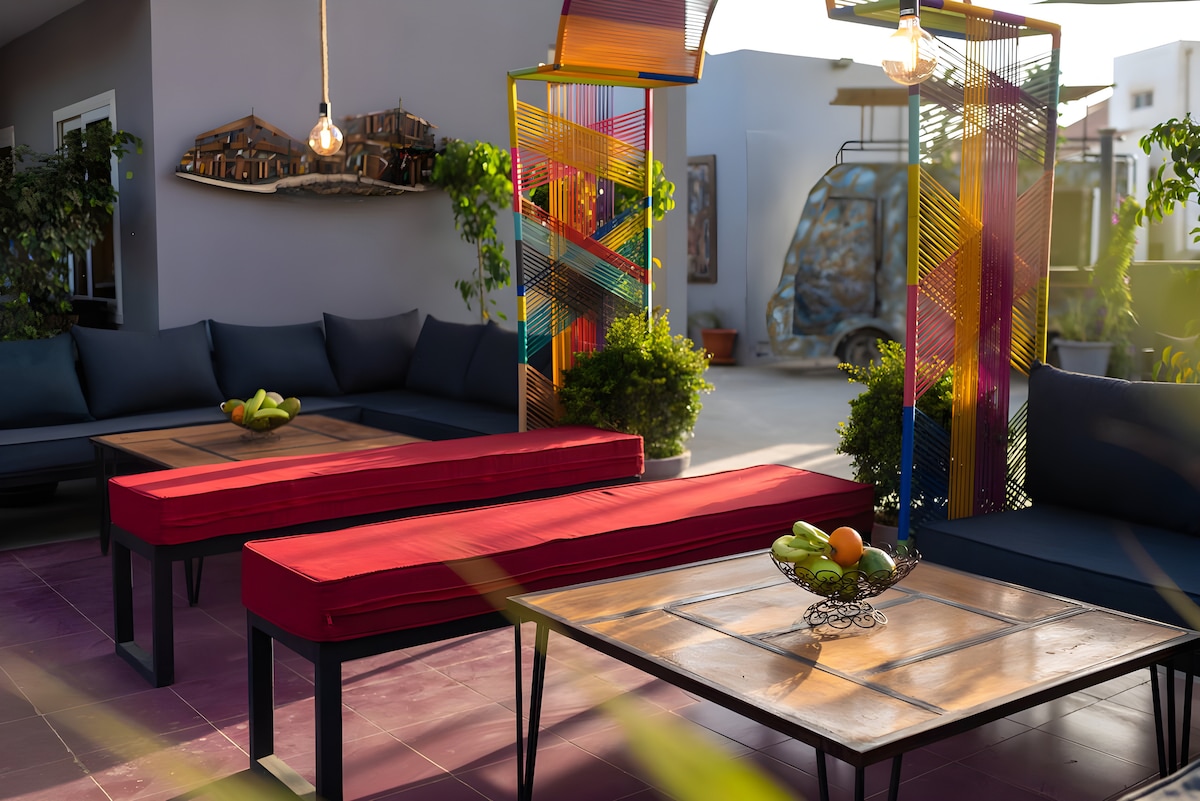 An outdoor lounge area is presented with modern seating featuring dark blue cushions and red upholstery. Low wooden tables, styled with fruit arrangements, complement the vibrant decor, which includes colorful vertical structures and surrounding greenery, creating a relaxed and inviting environment.