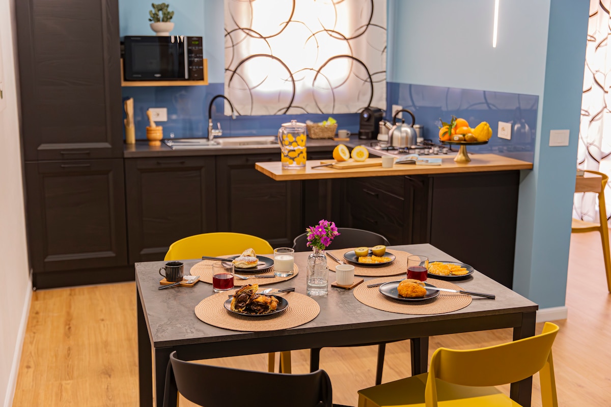 A colorful dining area is set with plates of pastries and drinks on a textured table. A modern kitchen features dark cabinetry, a microwave, and a kettle. Natural light enters through a decorative curtain, illuminating a vibrant fruit bowl on the counter.