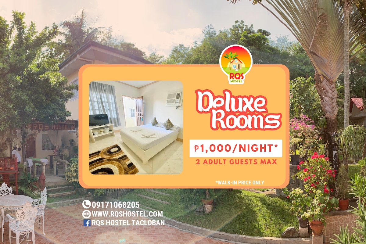 The image features a bright and airy Deluxe Room with expansive windows allowing natural light to fill the space. A spacious queen-sized bed is centered in the room, complemented by a light-colored bedding set. The room is visually enhanced by minimalist decor and a soft carpet underfoot.