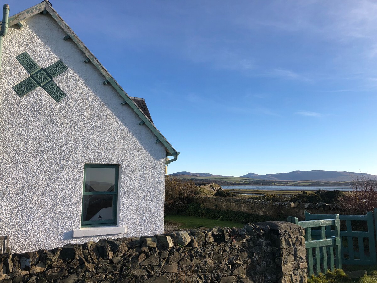 A charming white cottage is depicted with green accents on its exterior. A stone wall borders the property, while a wooden gate leads to a scenic view of the landscape beyond, showcasing open fields and distant hills under a clear blue sky.