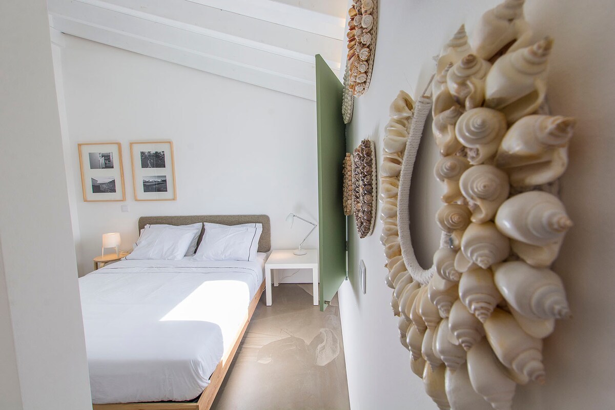 The spacious bedroom features a comfortable bed with crisp white linens. A round mirror adorned with seashells adds a decorative touch, while framed black-and-white photographs complement the pale walls. Soft natural light enters through the open space, creating a serene environment.