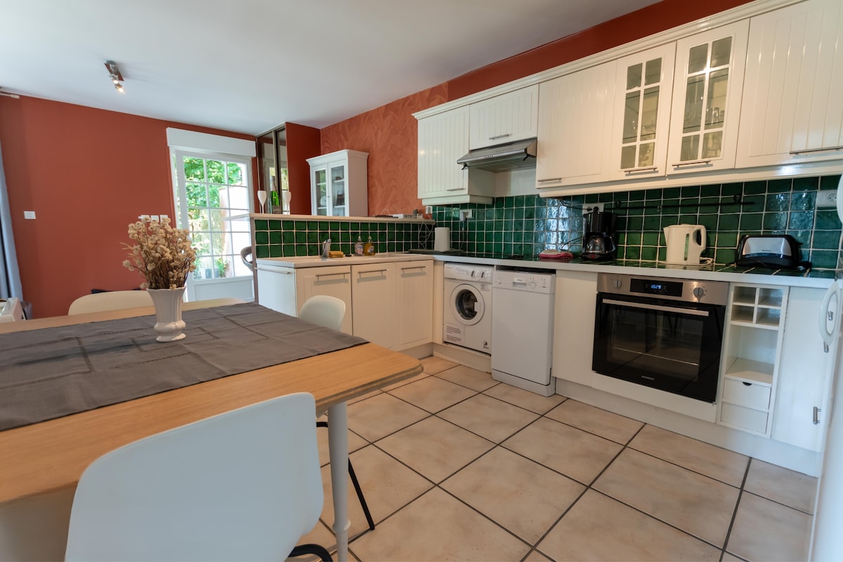 The kitchen features modern appliances set against vibrant green and white cabinetry. A wooden dining table is positioned centrally, surrounded by light-colored chairs. Large windows offer natural light, and a door leads to the outdoor area, enhancing the space's openness.