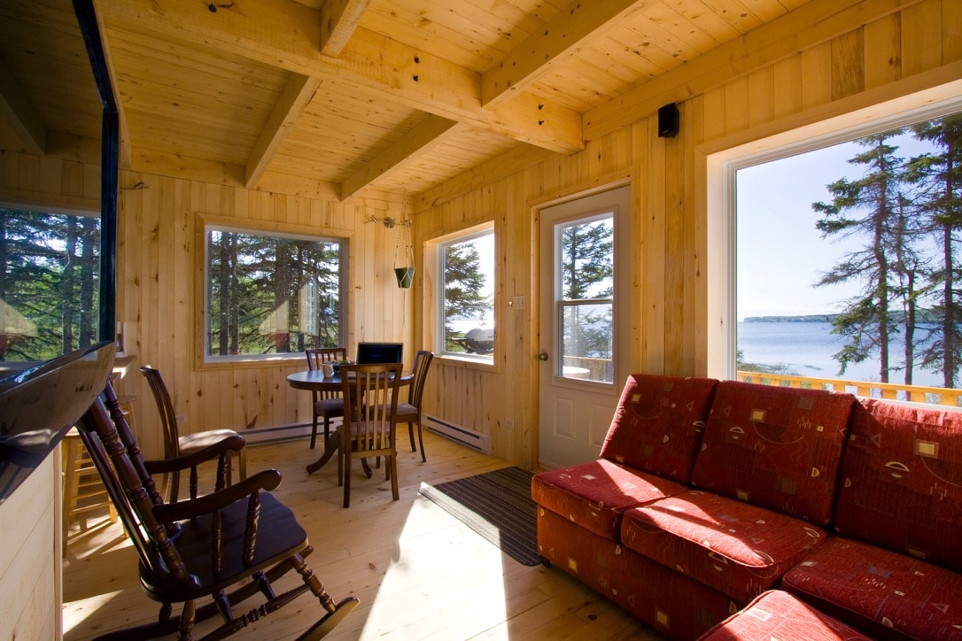 A light-filled living space is characterized by wooden beams and panoramic windows, offering views of the water and surrounding trees. A cozy couch and rocking chair are positioned beside a round dining table, creating an inviting area for relaxation and meals.