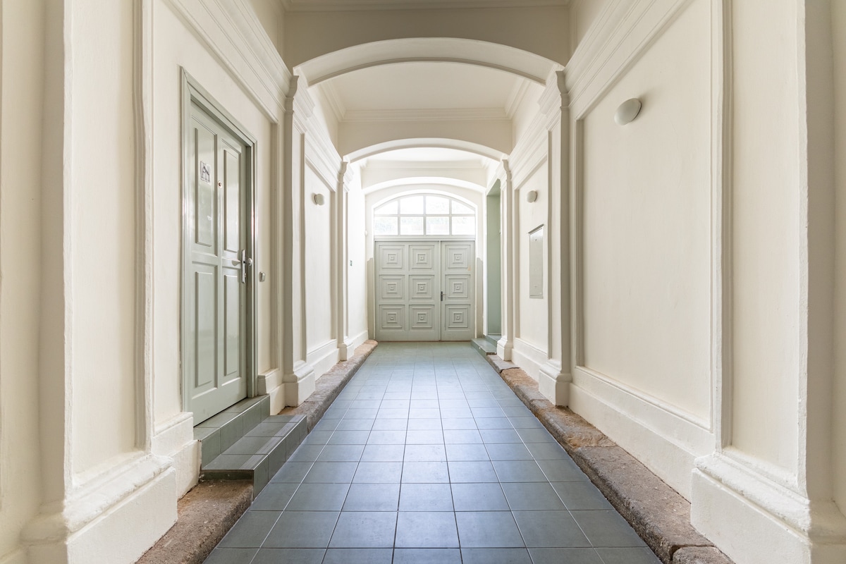 A spacious corridor leads to the apartment, featuring neutral walls and elegant archways. The entrance door is visible alongside a light-colored wooden door. An elevator is located at the end of the hallway, providing easy access to the apartment on the second floor.