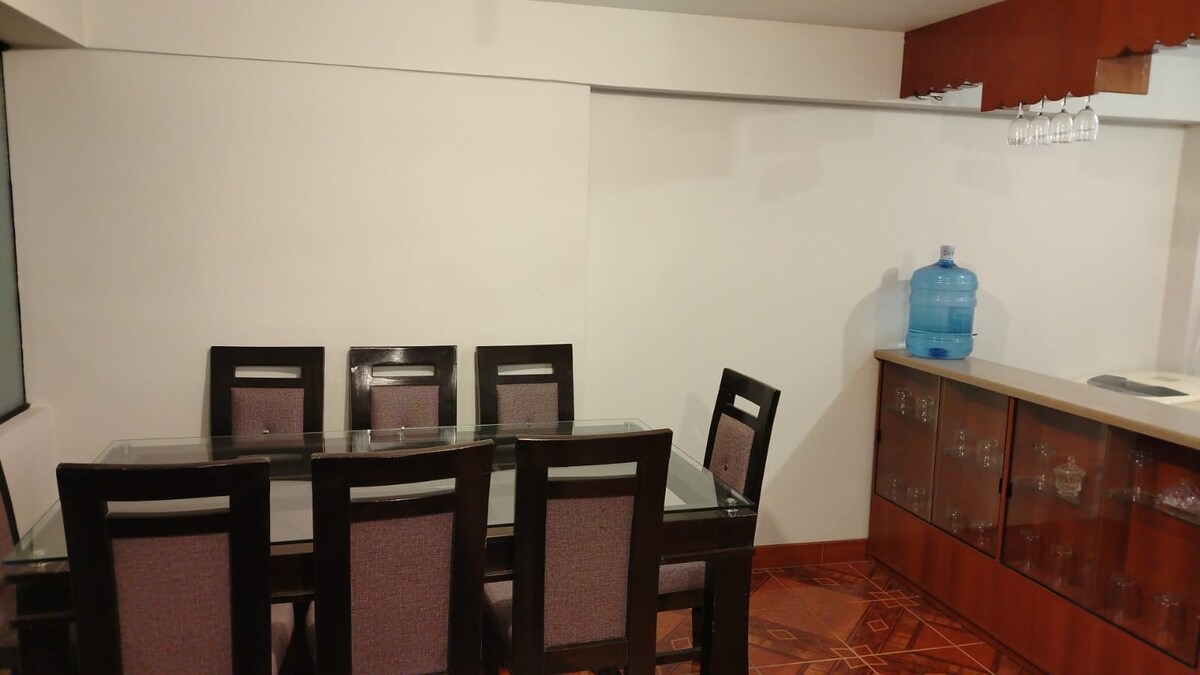A dining area is presented, featuring a glass-topped table surrounded by six dark wooden chairs with upholstered backs. The space is well-lit and includes a countertop with glass cabinets, a water dispenser, and a section for hanging glassware on display.