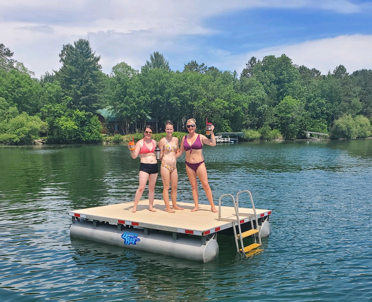 A floating platform in the water showcases three individuals posing cheerfully. Surrounding trees provide a lush green backdrop, with calm waters reflecting the sky. A ladder leads from the platform into the lake, creating an inviting space for relaxation and summer fun.