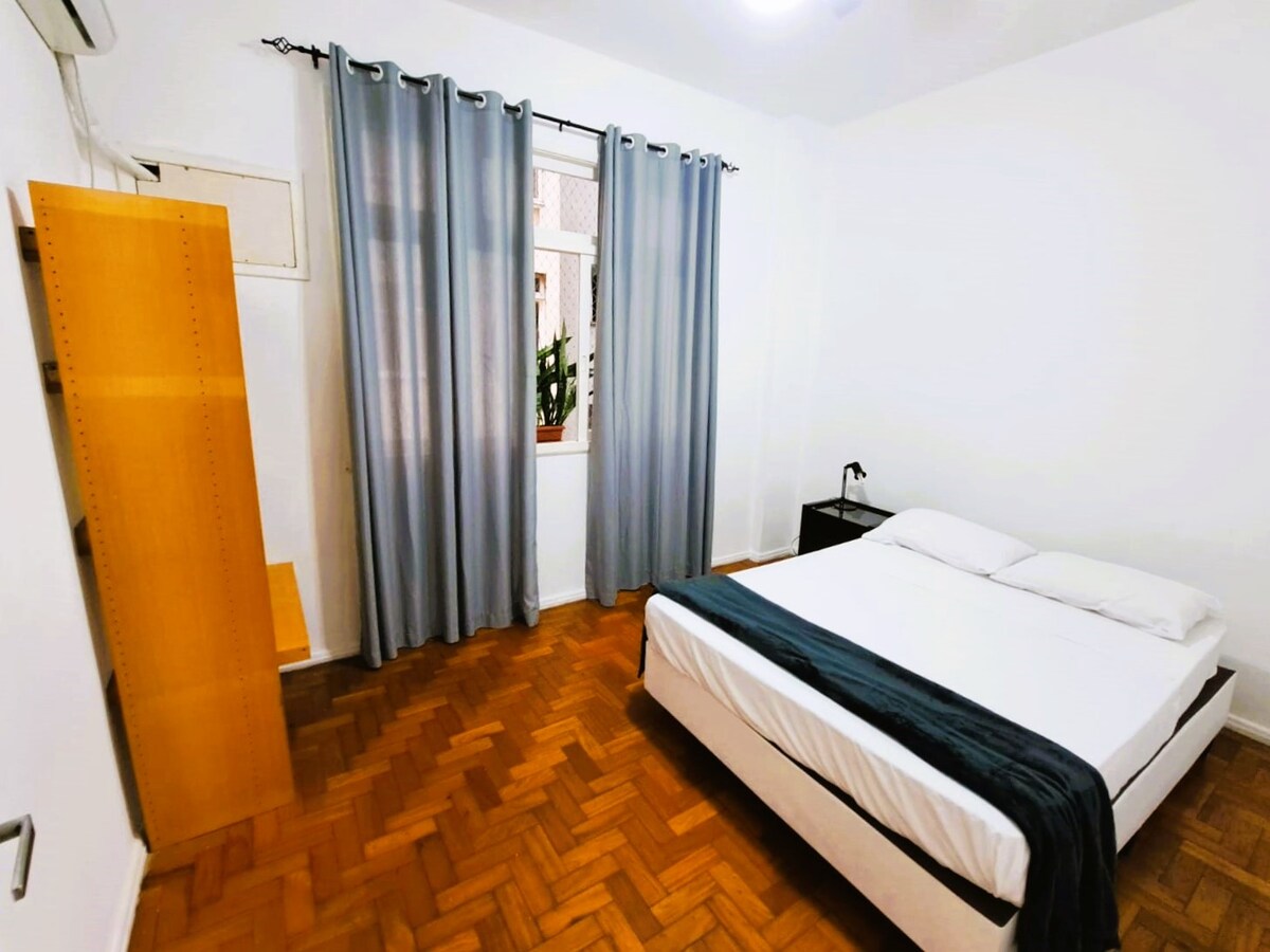 Quiet room with double bed, bedside table with light, open closet for clothes and towels, ceiling fan and air conditioner.