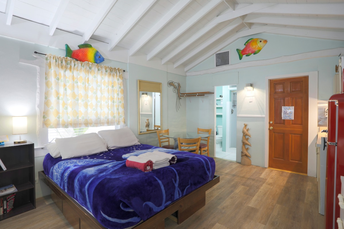 The interior space features a cozy queen size bed with fresh linens and colorful towels. Brightly colored fish decorations adorn the walls, complementing the soft yellow drapes above a window. A small dining area is visible, and a doorway leads to the adjoining bathroom.