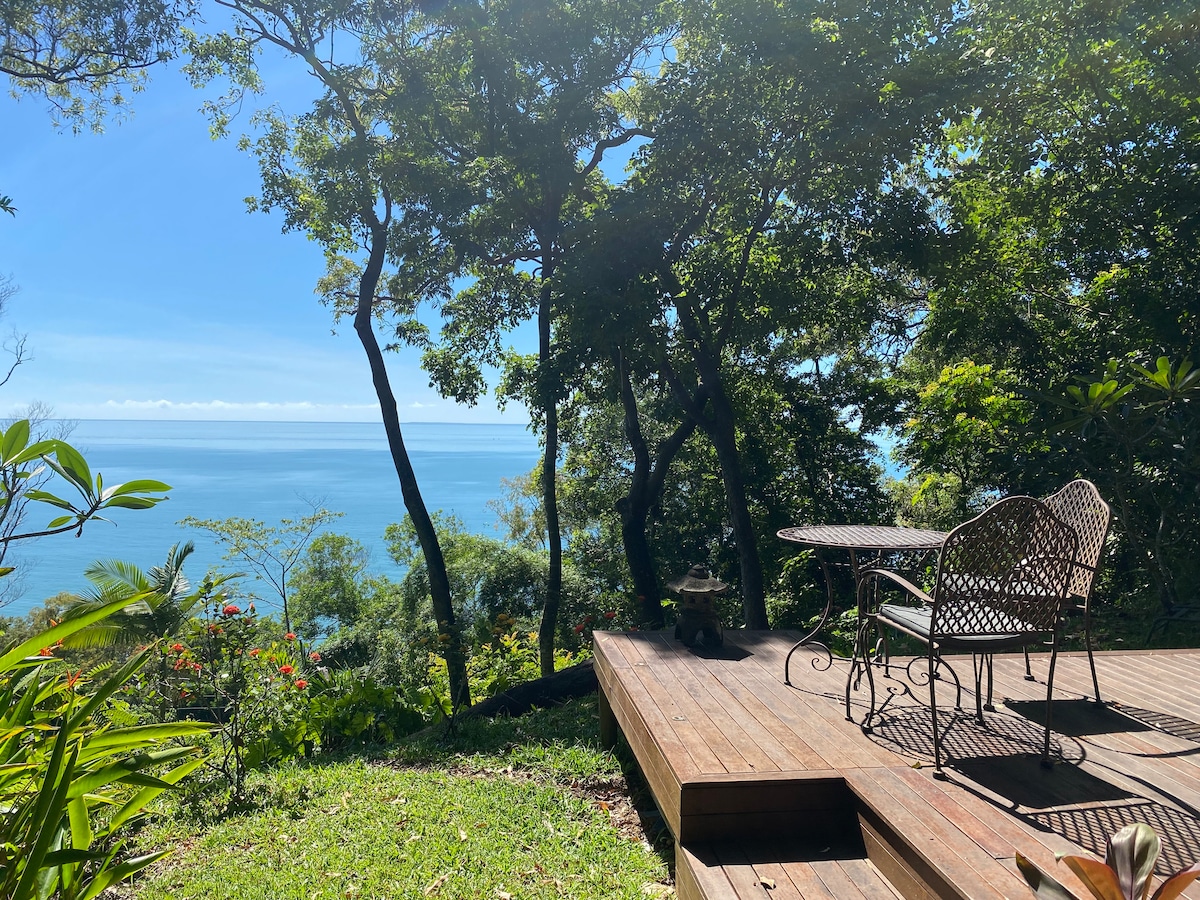 A wooden deck is surrounded by lush greenery and trees, offering a clear view of the ocean. A wrought-iron table and chairs are positioned on the deck, providing a serene spot to enjoy the natural beauty of the landscape.