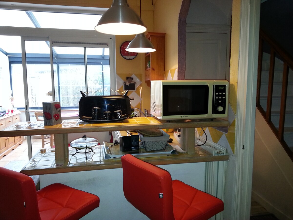A cozy kitchen area features modern appliances, including a microwave and a grill, positioned on a small counter. Two red bar stools are placed at the counter, inviting casual dining. Natural light enters through the adjoining glass-walled room, creating a bright atmosphere.