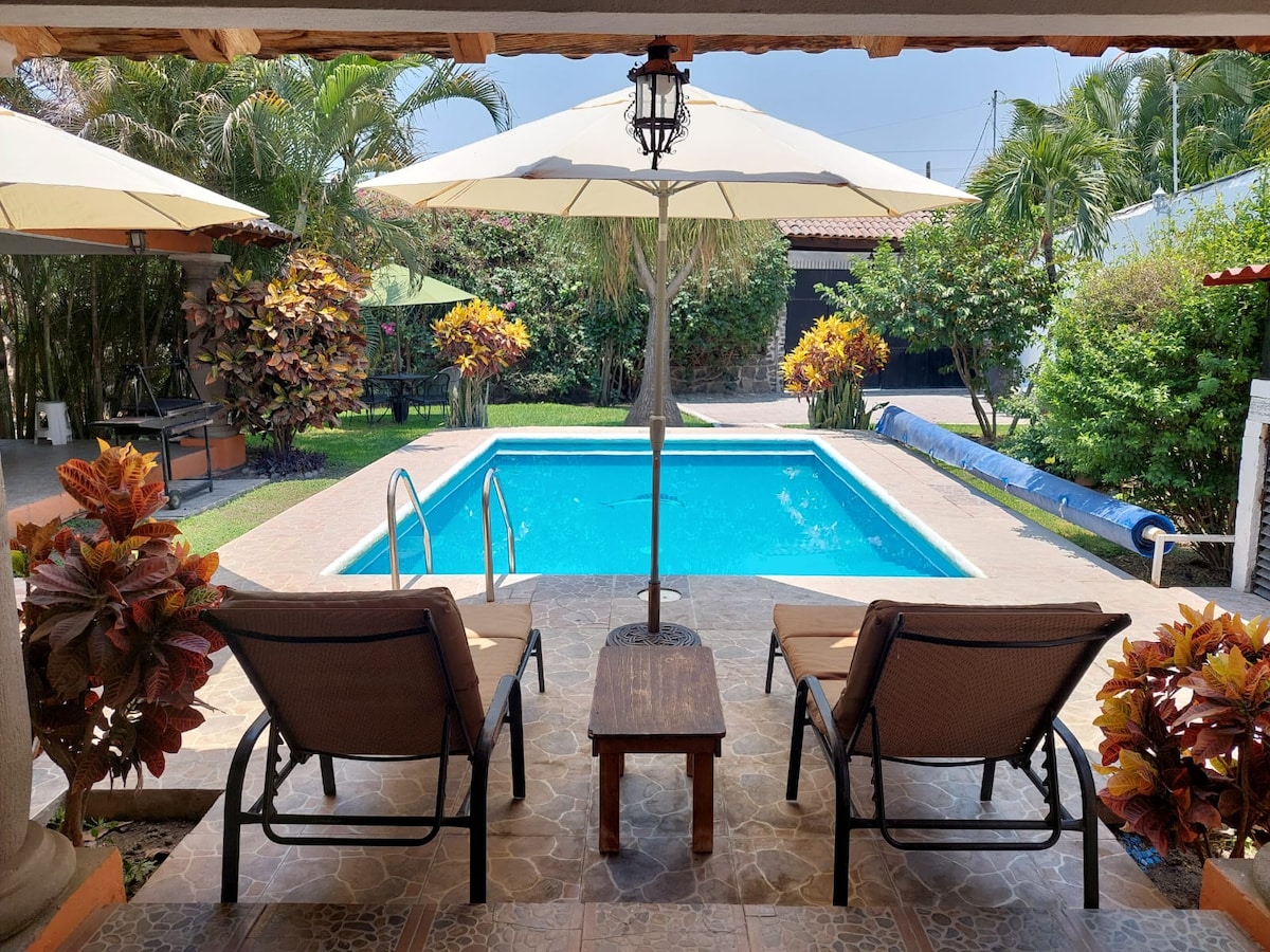 A view from a terrace features two lounge chairs positioned near a sparkling pool, surrounded by lush tropical plants and colorful flowers. Large umbrellas provide shade, and a small table sits between the chairs, inviting relaxation.