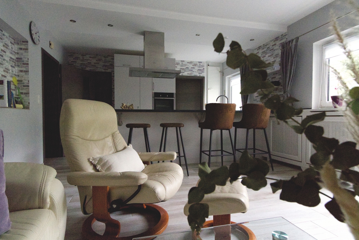 A modern living space features a comfortable beige armchair and wooden rocking chair. A sleek kitchen with a dining area is visible, showcasing high bar stools and contemporary cabinetry. Natural light enters through the window, enhancing the warm, inviting ambiance of the area.
