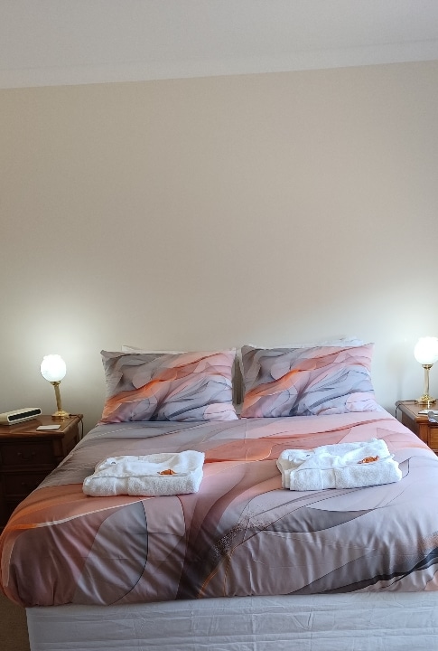 A spacious bed is centered in the room, adorned with soft bedding featuring gentle patterns. Two bedside lamps provide a warm glow, and fresh towels are neatly arranged at the foot of the bed. Neutral walls create a calming ambiance.