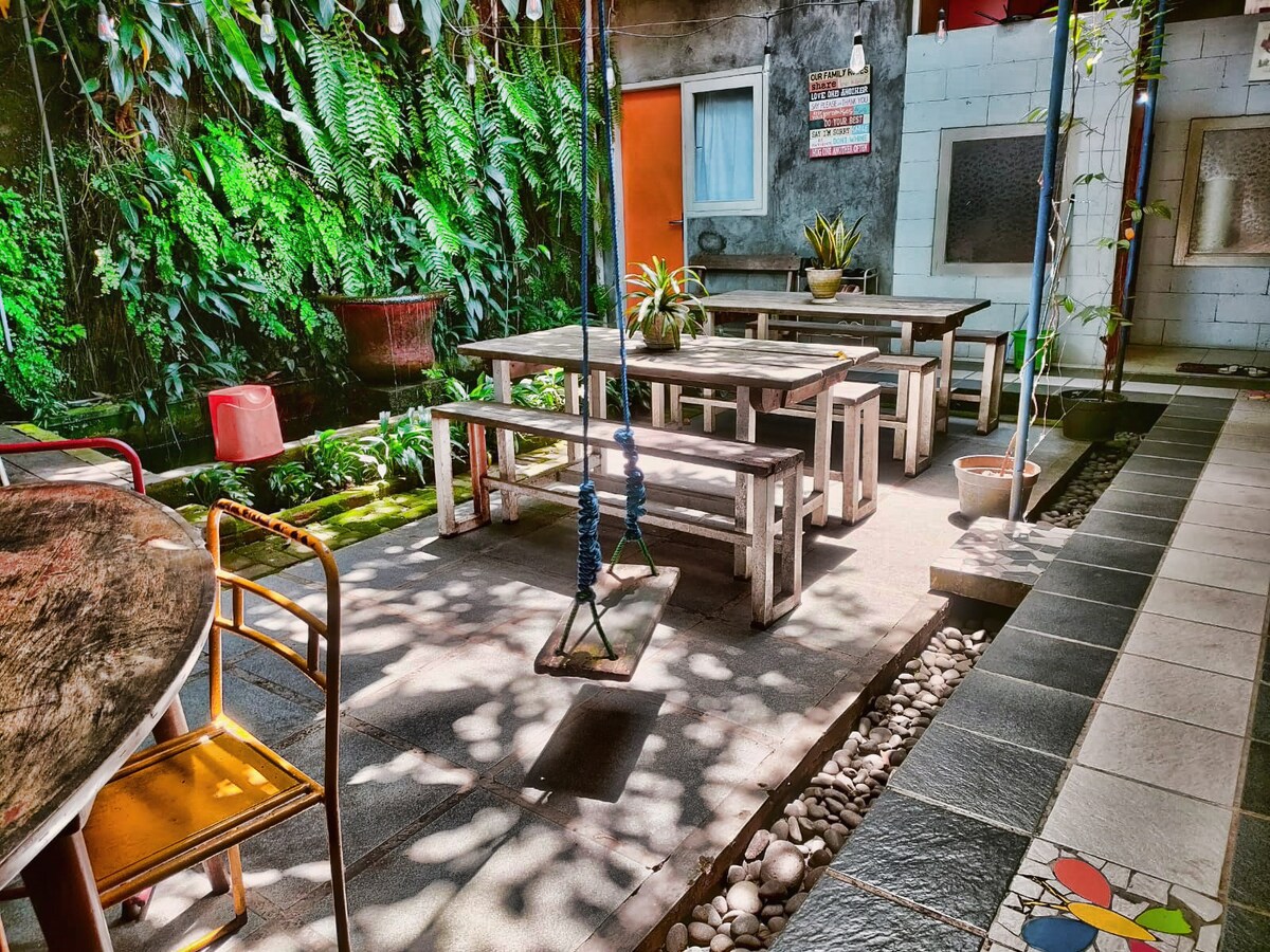 An outdoor communal area is featured, showcasing several wooden tables and benches set amidst greenery. Sunlight filters through leaves, creating patterns on the floor. A swing hangs from a beam, surrounded by potted plants and decorative stones, inviting communal gatherings in a serene environment.