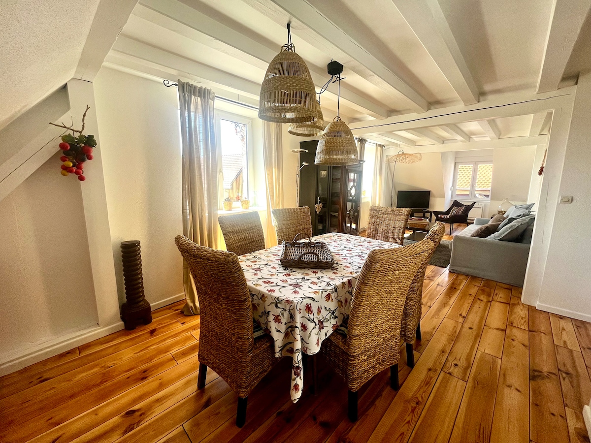 A dining area features a wooden table surrounded by six woven chairs, all set against a backdrop of wooden beams and warm-toned floorboards. Natural light filters through the windows, enhancing the space, while decorative pendant lights add subtle illumination.