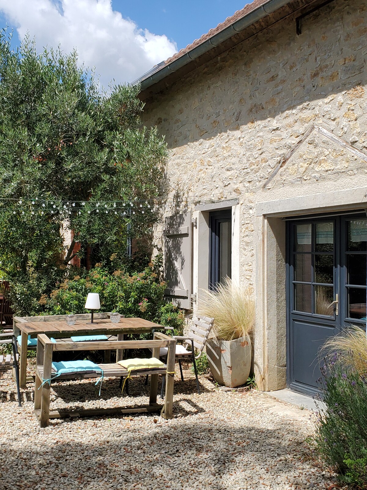 A charming outdoor terrace features a rustic wooden table surrounded by simple chairs, offering a peaceful setting. Lush greenery and decorative plants enhance the inviting environment, while stone walls and large windows reflect the character of the restored building.