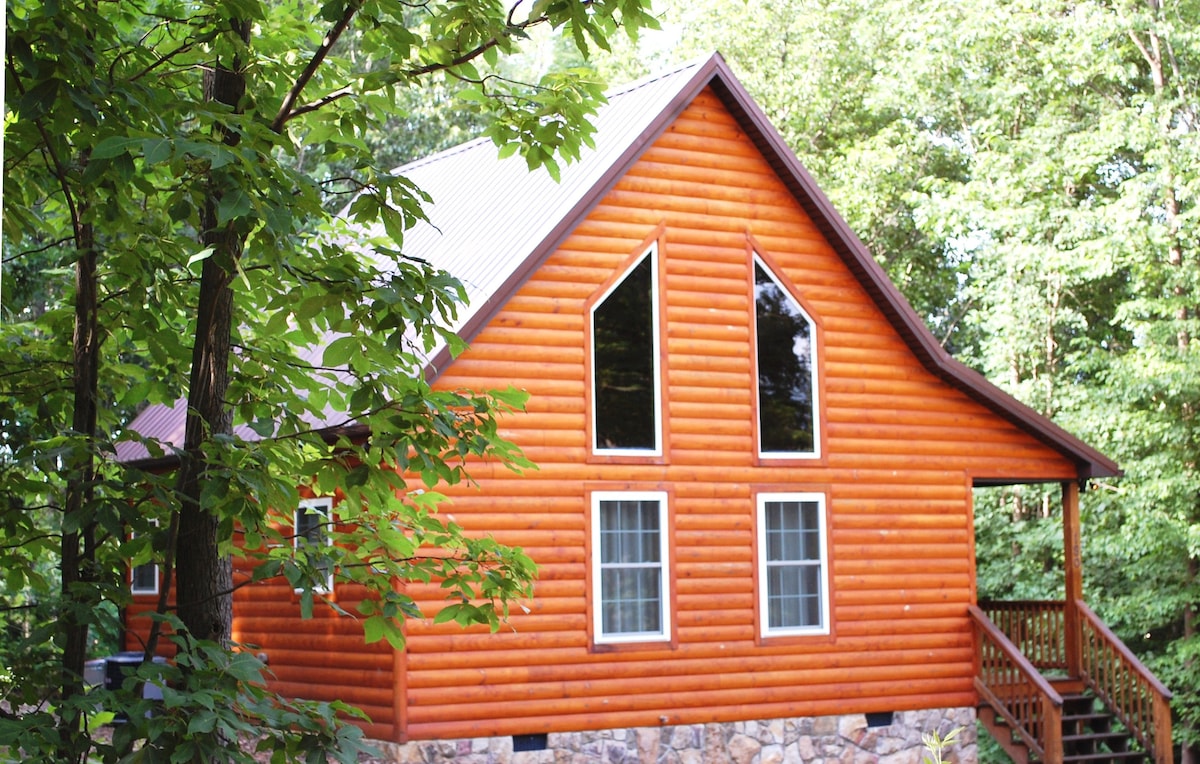 The cabin is framed by lush greenery, showcasing a striking log exterior in warm hues. Large triangular windows provide ample natural light, complemented by traditional double windows on either side. A wooden deck leads to the front entrance, enhancing the inviting appeal of the home.
